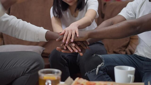 Handstack of unrecognizable African American men and Asian woman. Group of friends meeting indoors sitting on comfortable couch stacking hands