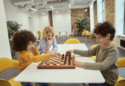 Competition. Joyful little diverse boys sitting at the table and playing chess in school