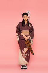 Japanese woman with traditional hairdo and kimono holding umbrella on pink background