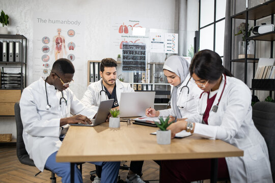 Four Focused Multiethnic Doctors Having Conference At Hospital Boardroom. Male And Female Heath Care Workers Using Modern Laptops At Tablets During Meeting.