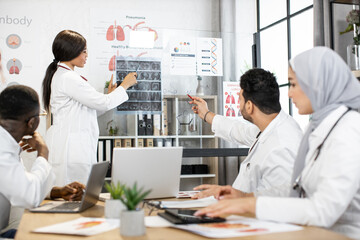 Group of four multiracial doctors using modern gadgets while examining tomography of patient during consilium. International colleagues sitting at room discussing various way of treatment.