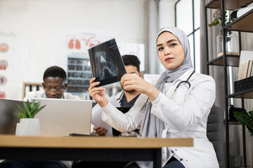 Fototapeta na wymiar Team of international competent doctors discussing results of x ray scan. Multiethnic medical workers sitting at conference room and using laptops with tablets. Muslim lady in hijab looks at the x-ray