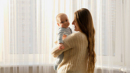 Happy smiling young mother with cute little baby standing at big window with curtains. Concept of family happiness and parenting