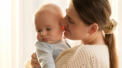 Portrait of smiling happy mother kissing her little baby son in cheeks. Concept of family happiness and parenting