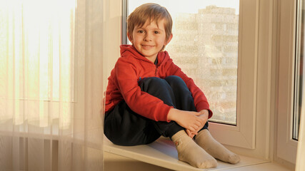 Naklejka premium Portrait of smiling little boy sitting on windowsill and looking in camera. COncept of staying at home and covid-19 pandemic