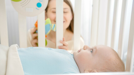 Portrait of young mother looking through wooden cradle banister on her little baby lying in cradle. Concept of parenting, family happiness and baby development