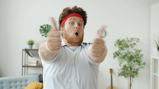 Portrait of crazy guy in sportswear showing thumbs-up hand gesture and making funny face indoors at home. Sports and overweight people concept.