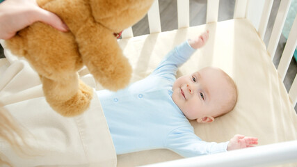 Mother giving teddy bear to her little baby boy lying in bed. Concept of parenting, family happiness and baby development