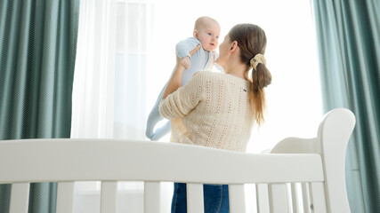 Young mother standing at cradle and holding her smiling baby son. Concept of parenting, family happiness and baby development