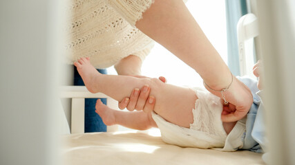 Mother changing diapers to her baby in cradle and kissing his little feet. Concept of parenting, family happiness and baby development