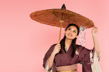 Fototapeta premium Happy japanese woman in traditional clothes standing under umbrella isolated on pink