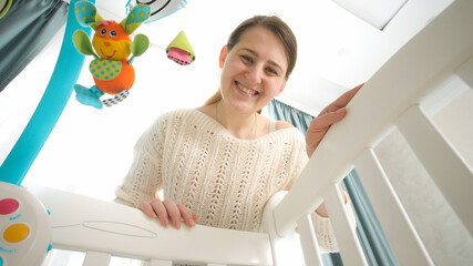 Happy smiling mother rocking cradle and looking at her sleeping little baby. Concept of parenting,...
