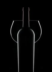 Closeup silhouette of a bottle of red wine in front of an empty wineglass over a black background.