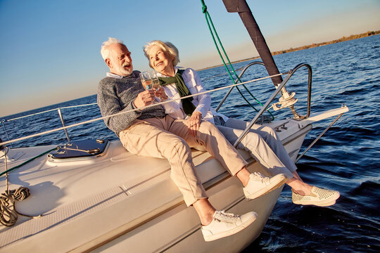 Happy Senior Couple In Love Drinking Wine Or Champagne And Laughing While Celebrating Wedding Anniversary On A Sailboat Or Yacht Deck Floating In A Calm Blue Sea