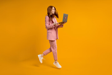 Red haired woman run jump typing laptop wearing elegant pink suit isolated on yellow background in studio, hurry up. side view portrait of lady working on laptop. Copy space for advertisement.