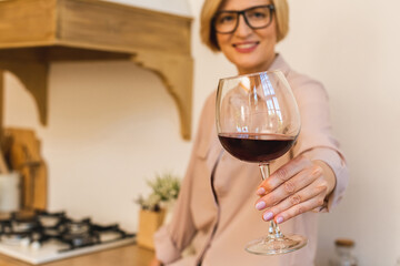 Happy mature senior elderly blonde woman in kitchen drinking red wine. Holding a glass with wine.
