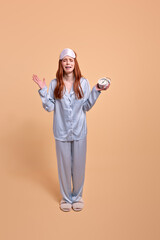 Crying Redhead Woman Want To Sleep In The Morning, Holding Clocks In Hands, Upset, Need To Wake Up, Wearing Blue Pajamas, Isolated On Beige Background, Portrait
