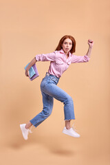 Fototapeta na wymiar Confused redhead female student rushing forwards with book in hands, hurry to university, wearing casual outfit, running, 1 September exam, isolated in studio, full-length portrait