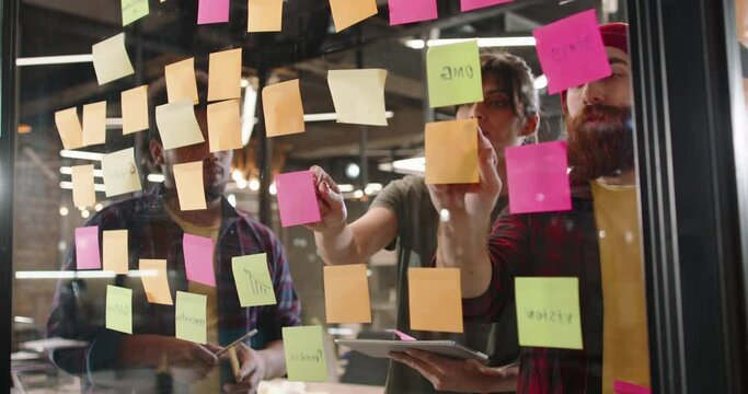 Creative multiethnic team of developers brainstorming ideas, putting stickers on glass board. Students working on project 4k footage