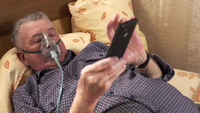 Old man is lying on the bed wearing an oxygen mask and watching the mobile phone in his hand. Elderly ill person is resting in a room. He has a chronic obstructive pulmonary disease. Theme of health.