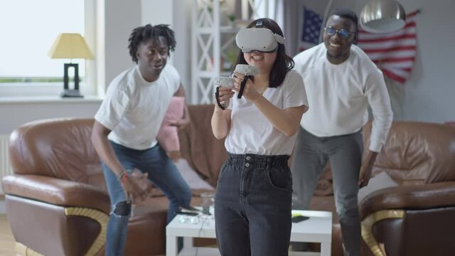 Young smiling Asian woman in VR headset boxing in augmented reality as African American men cheering supporting friend. Happy carefree young people resting gaming indoors on weekend - Powered by Adobe