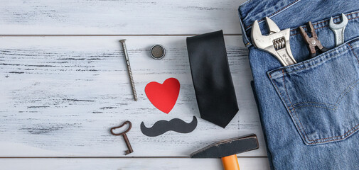 Construction tools and jeans on the table.
Construction tools, jeans, a tie and a paper heart with a mustache lie on the right on a white wooden table with space for text on the left, close-up top vie