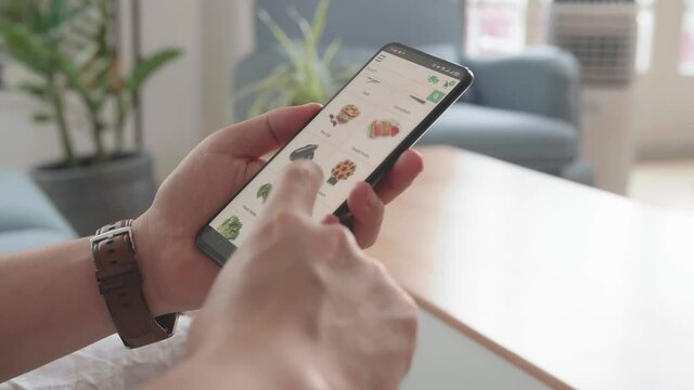 Man Sitting On Couch Using Smartphone Buys In Internet Shop. Male Selects Vegetables In Grocery Online Store
