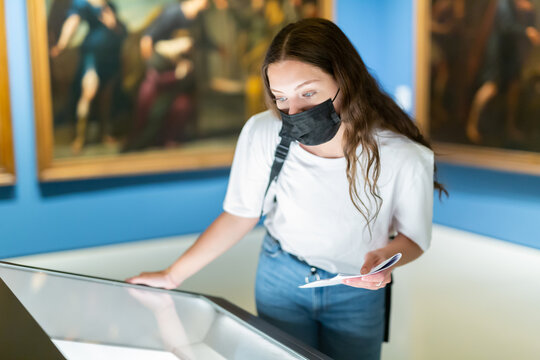 Focused young girl in a protective mask, visiting a museum during a pandemic, looks at the exposition behind a glass..case, holding an information booklet