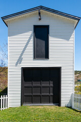 A tall double story white wooden clapboard siding building with a vintage wooden black garage door. There's a small black door above the structure. A white wooden picket fence encloses the garage.