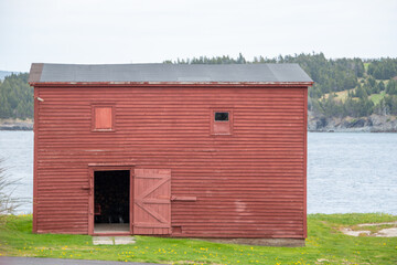 An old red barn of worn and weathered wood in a meadow surrounded by lush green grass. The barn has multiple windows. Behind the barn is a pond with trees and a mountain on the far side of the water. 