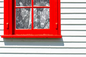 A bright red double hung vintage window with four glass panes with white lace curtains. The residential window is on the exterior of a narrow horizontal white wooden clapboard siding wall. 
