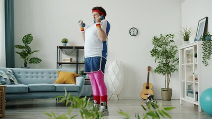 Crazy overweight sportsman in colorful outfit is training with dumbbells enjoying success indoors in apartment. Sporty youth and healthy activity concept.