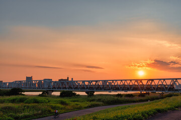 夕暮れの荒川河川敷。夕日をバックに橋を走る列車と自転車のある風景。