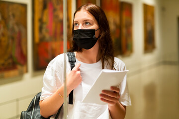 Focused girl wearing a protective mask, visiting an art museum during a pandemic, looks at an exhibit located behind..glass