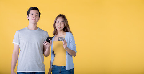 Asian couple holding credit card and smartphone while looking at a camera isolated on yellow background