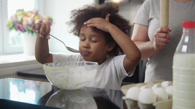Family baking - black little girl eating the rests of a liquid dough from the bowl