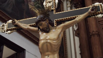 Medium shot in upward tilt motion showing a wooden statue of Jesus Christ crucified on a wooden cross with gilt edges, with the walls of the church out of focus behind. - Powered by Adobe