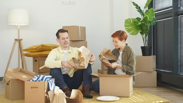 Delighted woman and husband unpack cardboard boxes with new tableware, carry plates, move into new apartment, look gladfully at each other, sitting on floor. Relocation concept