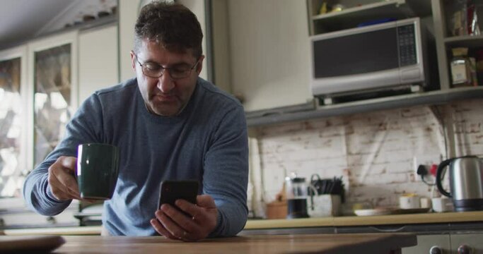 Smiling caucasian man using smartphone, drinking coffee in kitchen at home