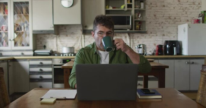 Caucasian man sitting at table, using laptop and drinking coffee at home