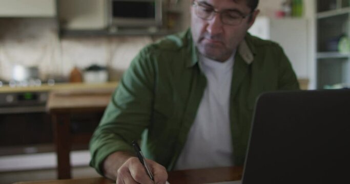 Caucasian man sitting at table, using laptop and making notes at home