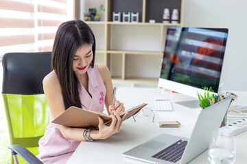 Beautiful Asian businesswoman sitting and taking notes with laptop at the office.