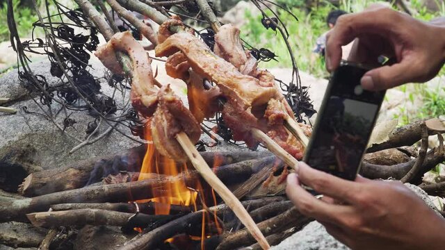Cooking meat on fire in the jungle with another guy making picture with his iphone. Sausages deliciously barbecued over open charcoal fire using carved wooden sticks. safe family hiking trip to forest
