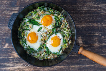 Shakshuka, fried eggs with green wild garlic leaves, onion, pepper and spices in cast iron pan, close up