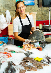 Female worker of fish shop in apron offering fresh raw flatfish for sale