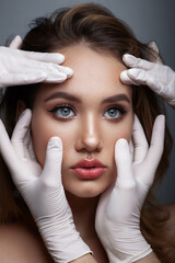 Close-up portrait of young female standing with naked skin, face is touched by doctors in gloves, preparing her for plastic surgery procedures, isolated on studio background