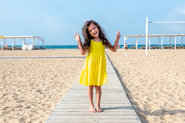 Adorable curly toddler girl in a yellow dress plays on the white sand beach 