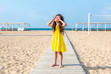 Adorable curly toddler girl in a yellow dress plays on the white sand beach 