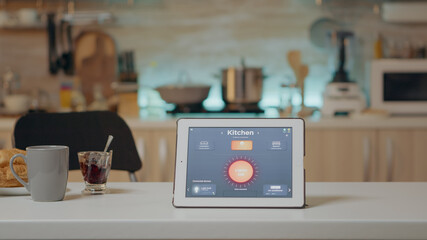 Tablet with intelligent software placed on table in kitchen with nobody in, controlling light with high tech application. Notepad with smart home app in empty house automation system