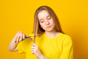 Sad and unhappy teen girl cutting her hair with scissors while standing on yellow background. Young student experiments with her hairstyle at home.